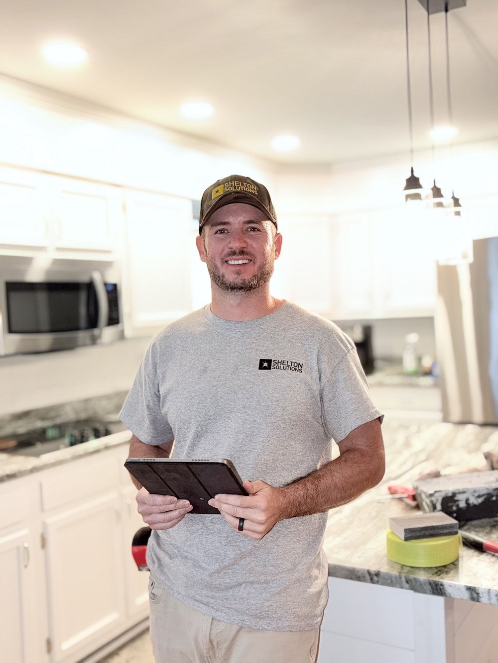 Josh Shelton in a Shelton Solutions cap and shirt, standing in a kitchen mid-remodel with a tablet, and a trowel and tape measure on the counter.